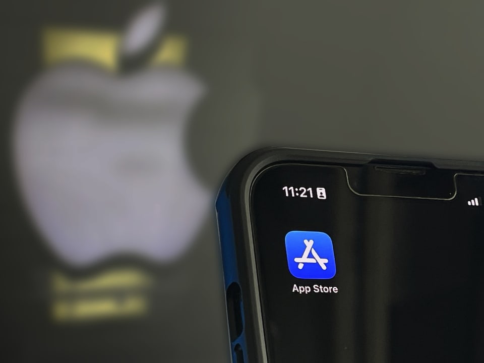 Auf einem iPhone-Bildschirm ist das Symbol des App Stores zu sehen. Im Hintergrund ist ein Apple-Logo unscharf.