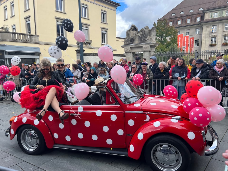 Parade mit rotem Auto, dekoriert mit weissen Punkten und Luftballons.