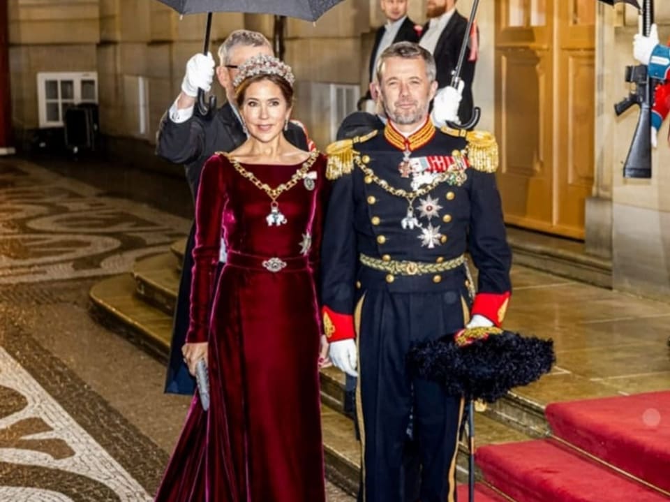 Eine Frau in einem edlen roten Kleid und ein Mann in königlicher Uniform auf einem roten Teppich.