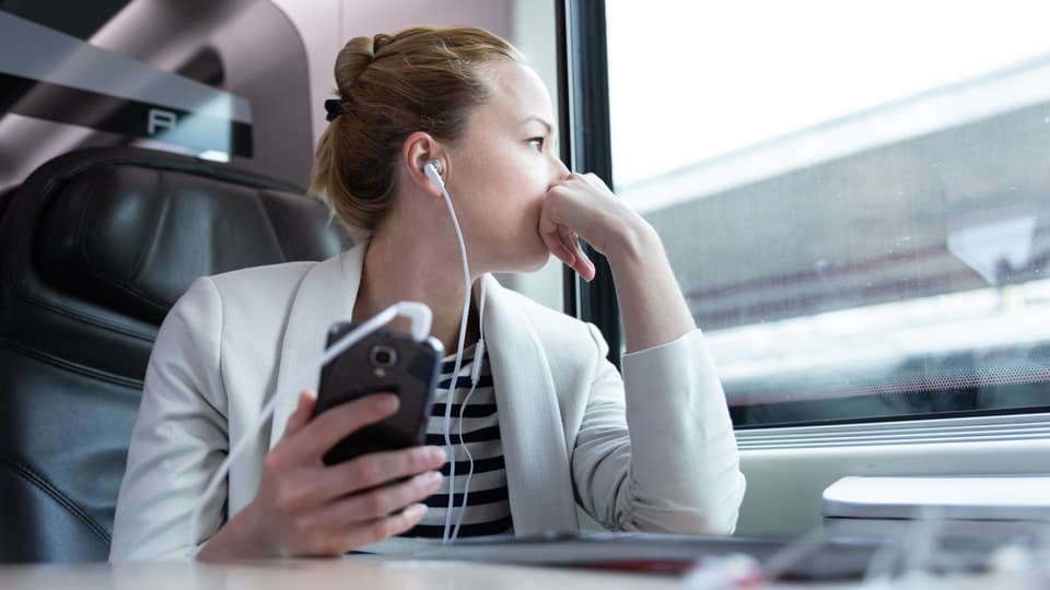 Frau sitzt im Zug, schaut aus dem Fenster und hört einen Podcast.