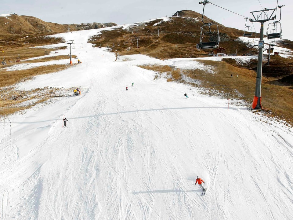 Skipiste in grün, eine Schneise ist beschniet und ein paar Wintersportler darauf unterwegs. 