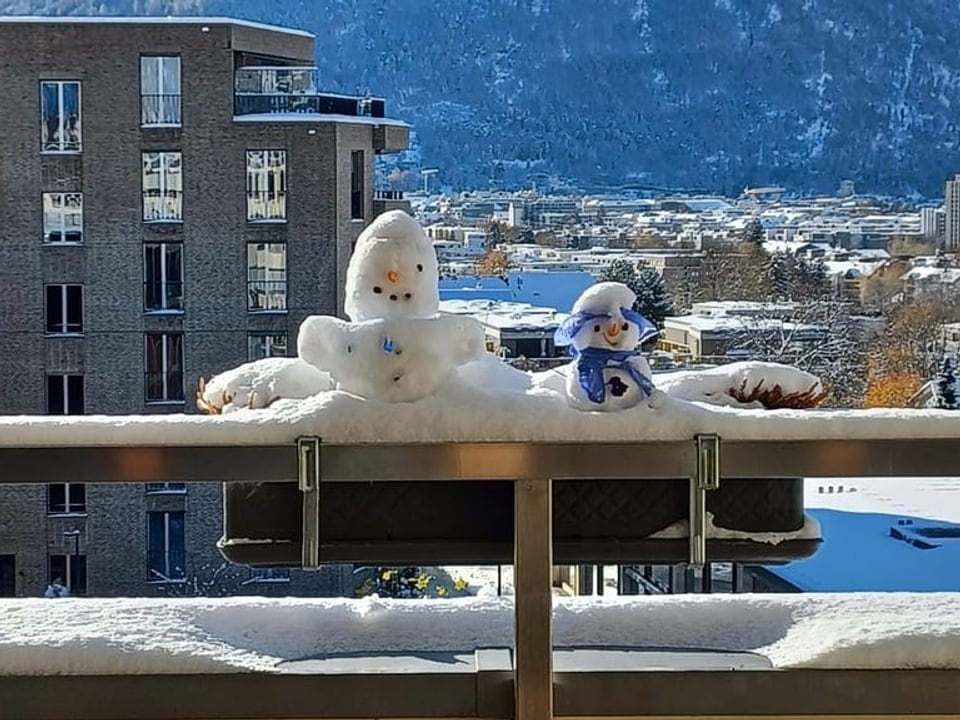 Schneefiguren auf Balkon mit Stadt und Bergen im Hintergrund.