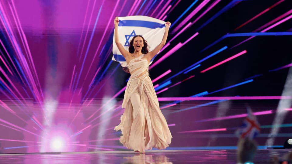 Frau in einem langen Kleid hält die israelische Flagge auf einer Bühne mit bunten Lichtstrahlen.