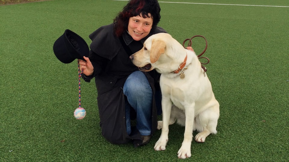 auf grünem Rasen kniet links eine Frau, hält einen Hut und einen Ball an einer Schnur, rechts sitzt der blonde Labrador.