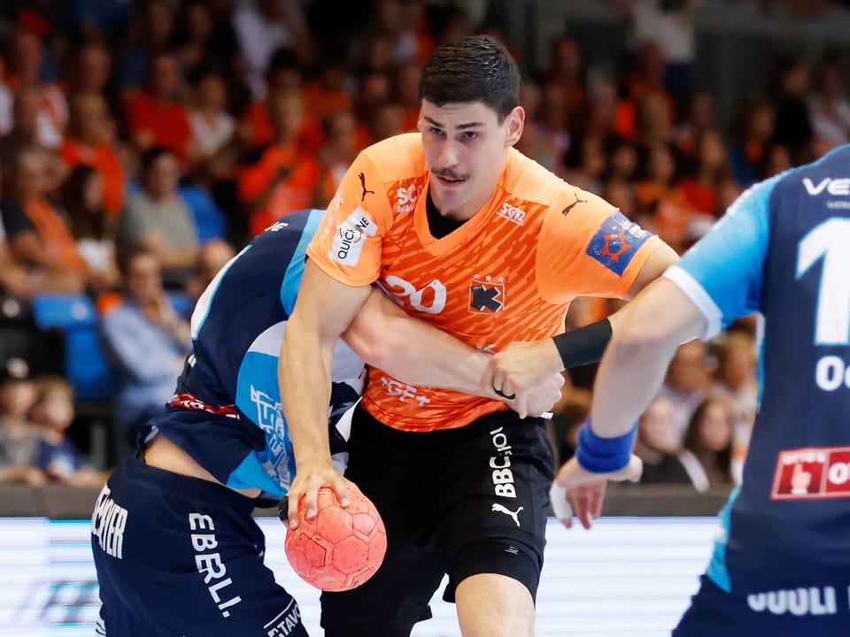 Handballspieler hält Ball während eines Spiels, Gegenspieler in blauen Trikots.