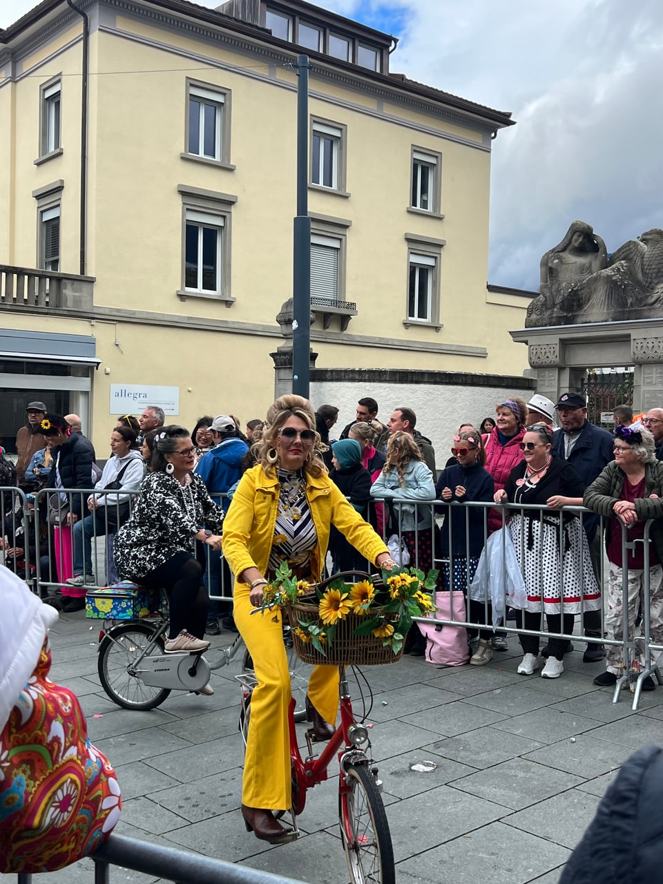 Frau in gelbem Anzug auf Fahrrad mit Sonnenblumenkorb bei Umzug.