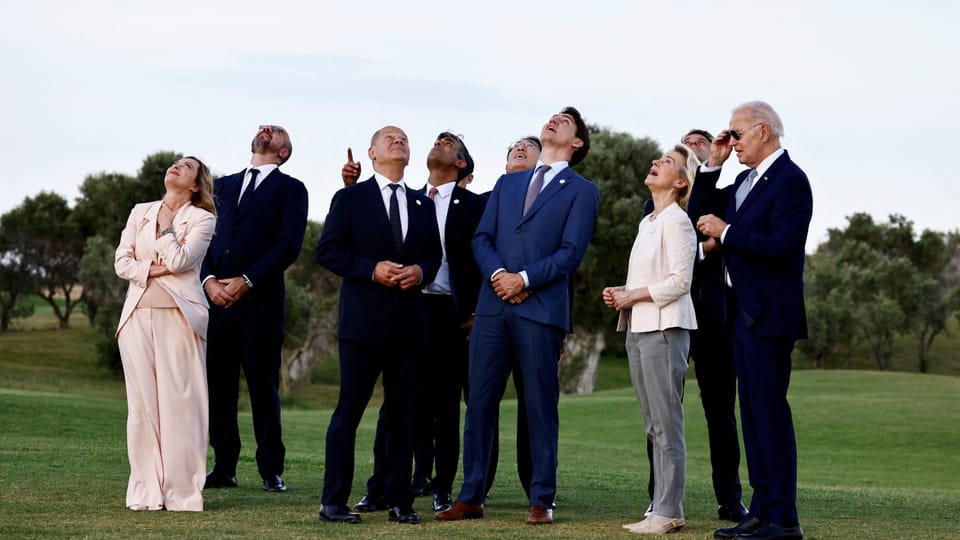 Acht Männer und Frauen in Anzügen blicken auf einer Wiese stehend gen Himmel