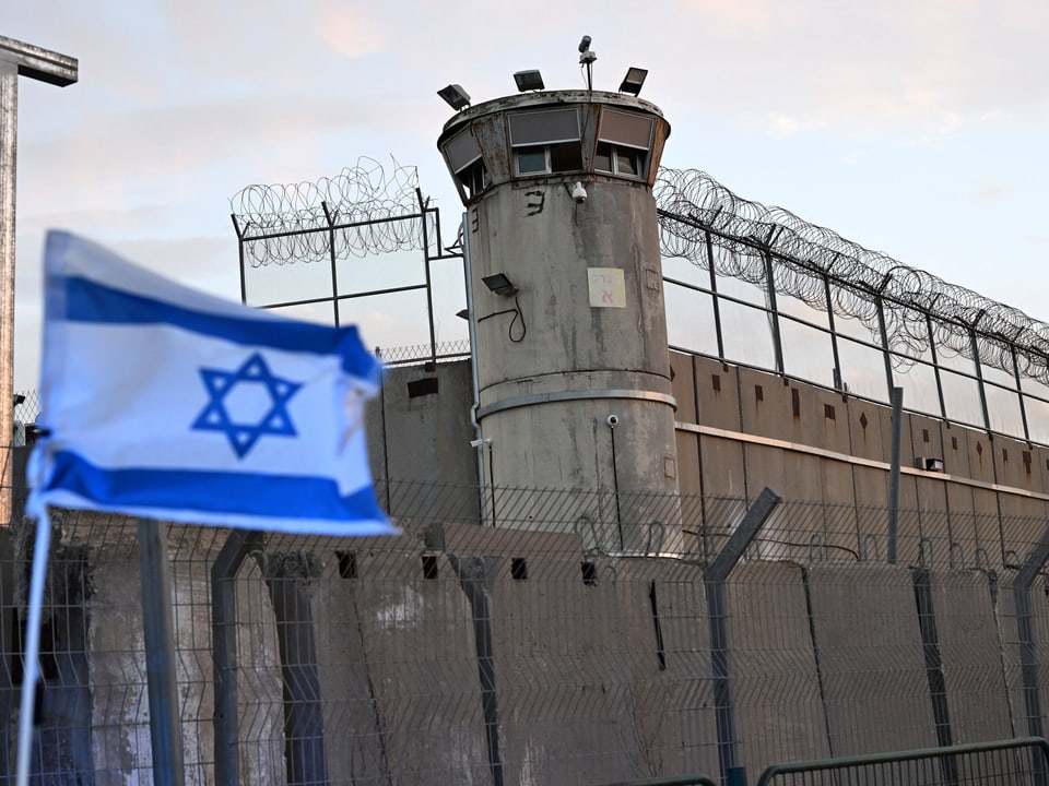 Wachturm hinter Stacheldrahtzaun mit israelischer Flagge im Vordergrund.