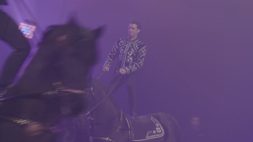 Bastian Baker steht auf einem galoppierenden Pferd und hält die Zügel in der Hand.