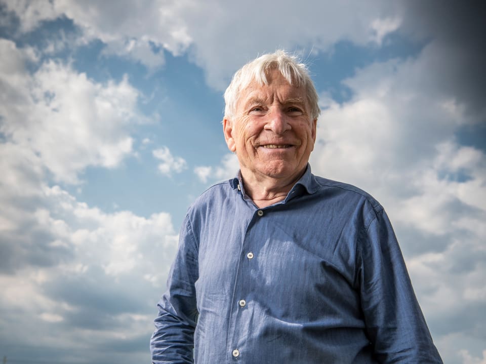 Älterer Mann in blauem Hemd lächelt vor wolkenbedecktem Himmel.