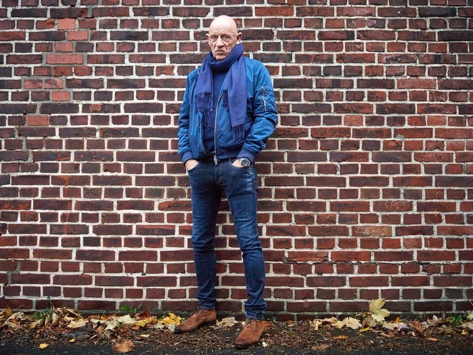 Älterer Mann in blauer Jacke und Schal vor Ziegelsteinmauer.