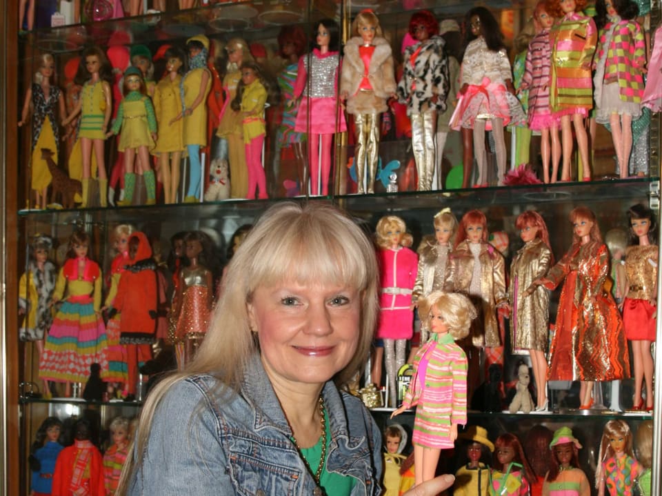 Eine Frau hält eine Barbie in der Hand. Hinter ihr steht ein Regal voller Puppen.