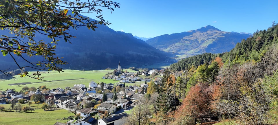 Blick auf ein Tal mit Dorf, Wiesen und Bergen im Hintergrund.