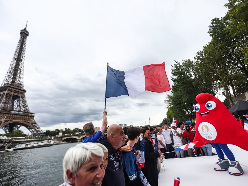 Menschen und ein Maskottchen feiern nahe dem Eiffelturm mit französischer Flagge.
