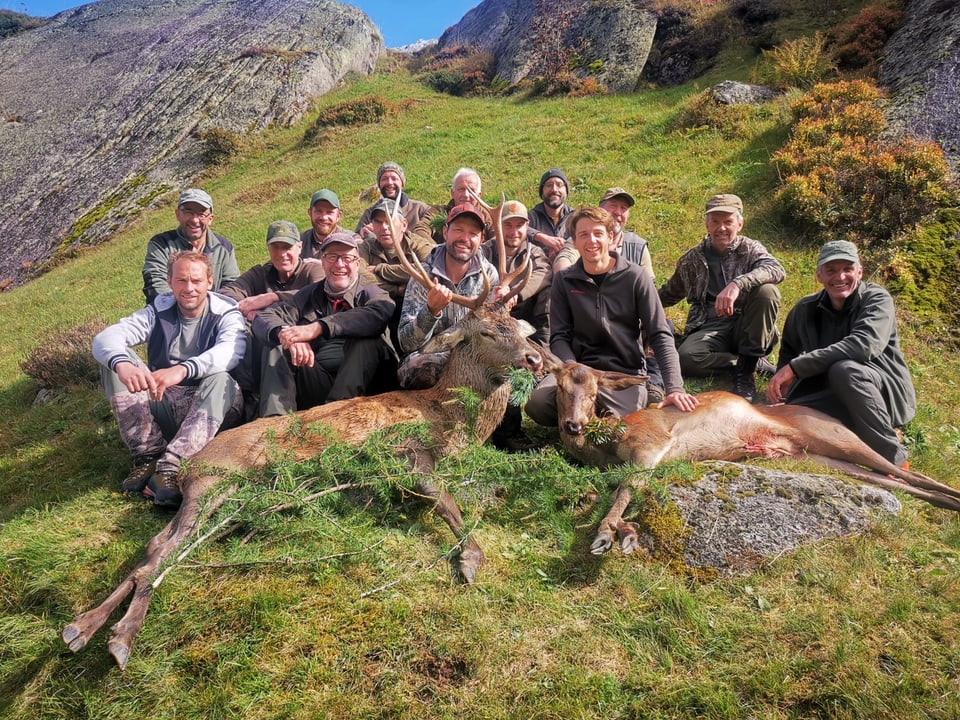 Gruppe von 15 Jägern mit zwei erlegten Hirschen auf einer Wiese vor Felsen.