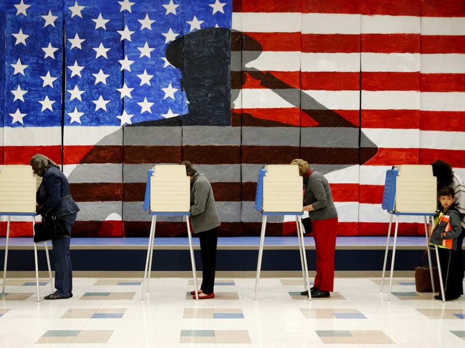 Menschen wählen in Wahlkabinen vor amerikanischer Flagge.