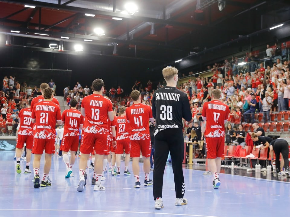 Handballmannschaft in roten Trikots geht auf das Spielfeld in einer Sporthalle.