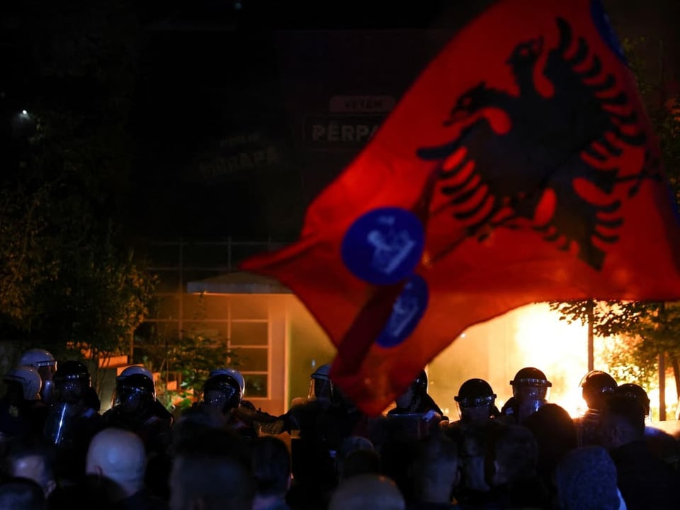 Albanische Flagge bei Nachtprotest, Menschenmenge und Feuer im Hintergrund.