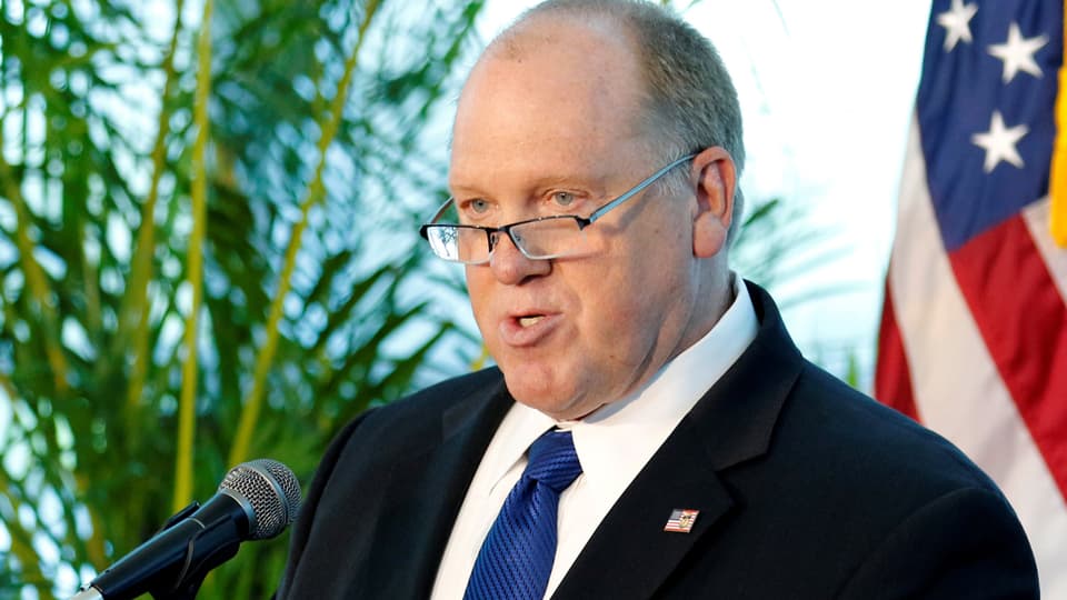 Mann mit Brille spricht vor Mikrofon, US-Flagge im Hintergrund.