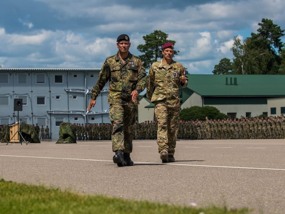 Zwei Soldaten in Uniform bei einer Militärparade, Soldatengruppen im Hintergrund.