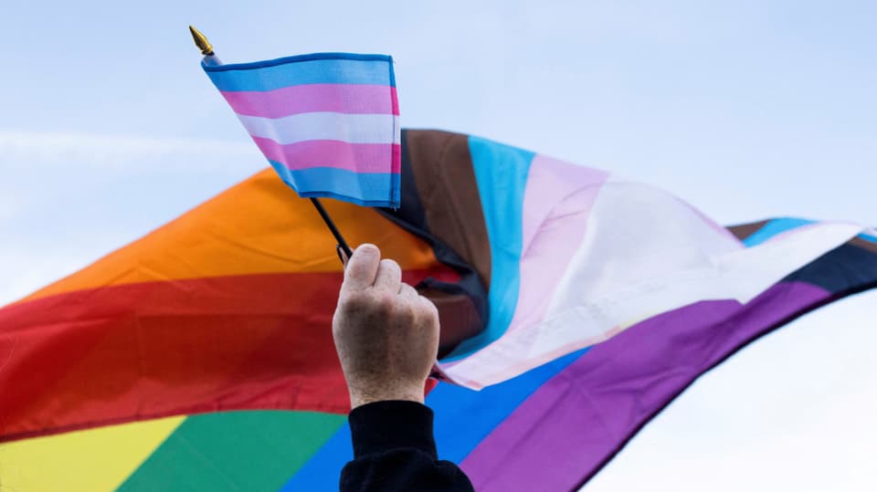 Eine handgehaltene Transgender-Flagge vor einer Regenbogenflagge.