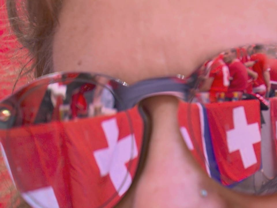 Schweizer Fans und Schweizer Fahnen spiegeln sich in der Sonnenbrille eines Mannes.