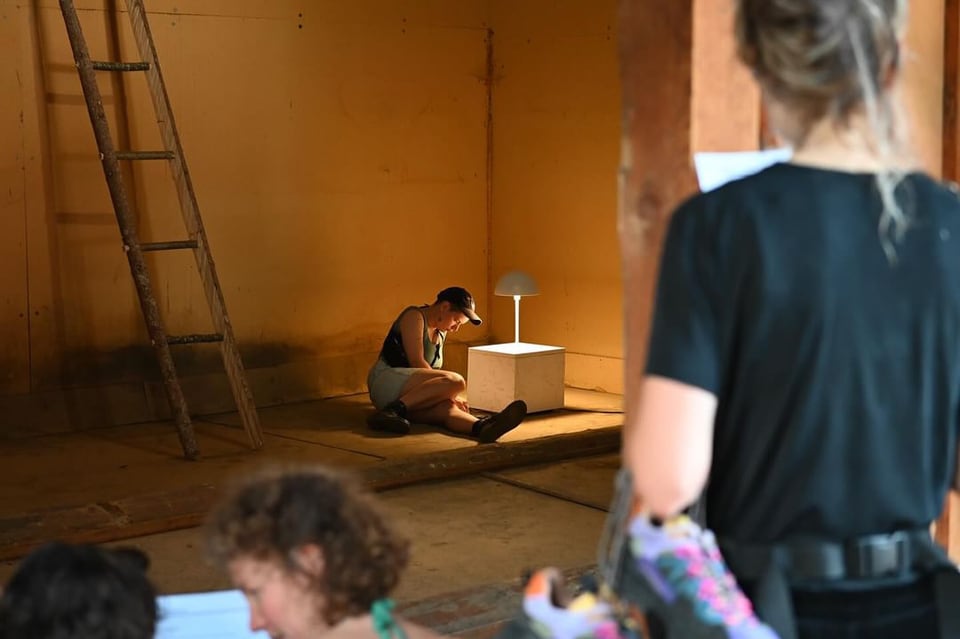 Frau sitzt am Buatsch Festival auf dem Boden neben einer Lampe in einem Atelier, andere Menschen im Vordergrund.