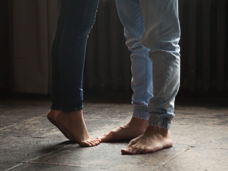 Beine und Füsse von Mann und Frau, wobei die Frau auf den Zehenspitzen steht, da sie viel kleiner ist.