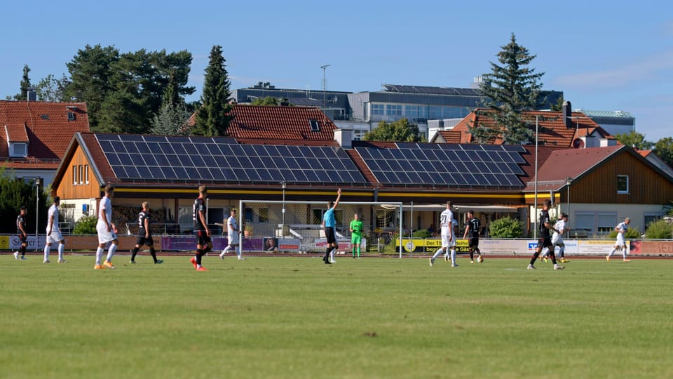 Ein Fissballfeld mit Spielern darauf, im Hintergrund ein Gebäude mit Solarpanelen.