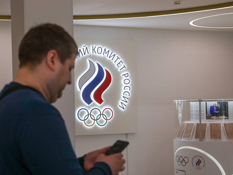 Mann mit Smartphone vor Logo des Russischen Olympischen Komitees.