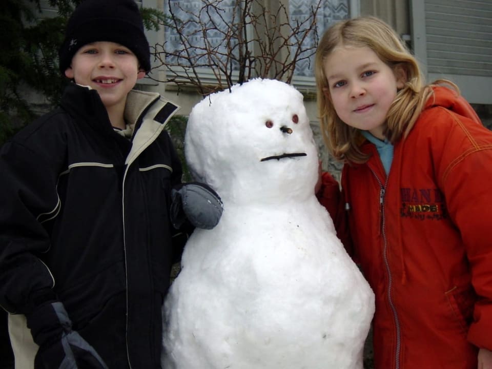Junge und Mädchen stehen neben einem Schneemann.