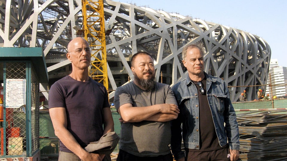 Drei Männer mittleren Alters stehen in legerer Kleidung auf einer Baustelle. Im Hintergrund zu sehen ein Stadion.