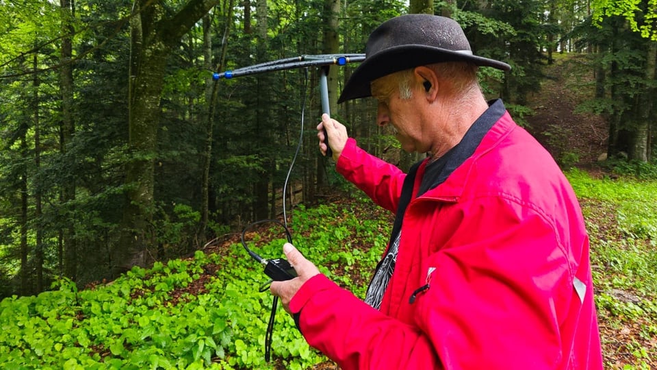 Ein Mann mit Roter Jacke steht am Waldrand und hat eine Art Antenne in der Hand.