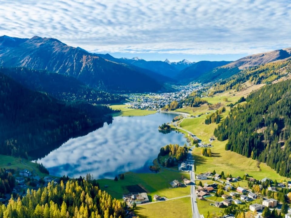 Landschaftsbild mit grünem Bergtal und See, in dem sich der bewölkt Himmel spiegelt. 