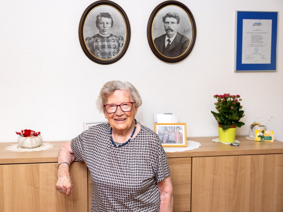 Marie Vogler-Gasser lehnt an ein Holzmöbel. An der Wand hängen zwei Schwarz-weiss-Fotos