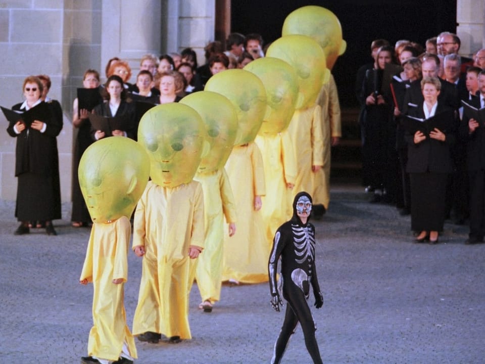 Eine Reihe von gelben Gestalten mit Alienmaske. Davor ein Mann in Skelettkostüm.