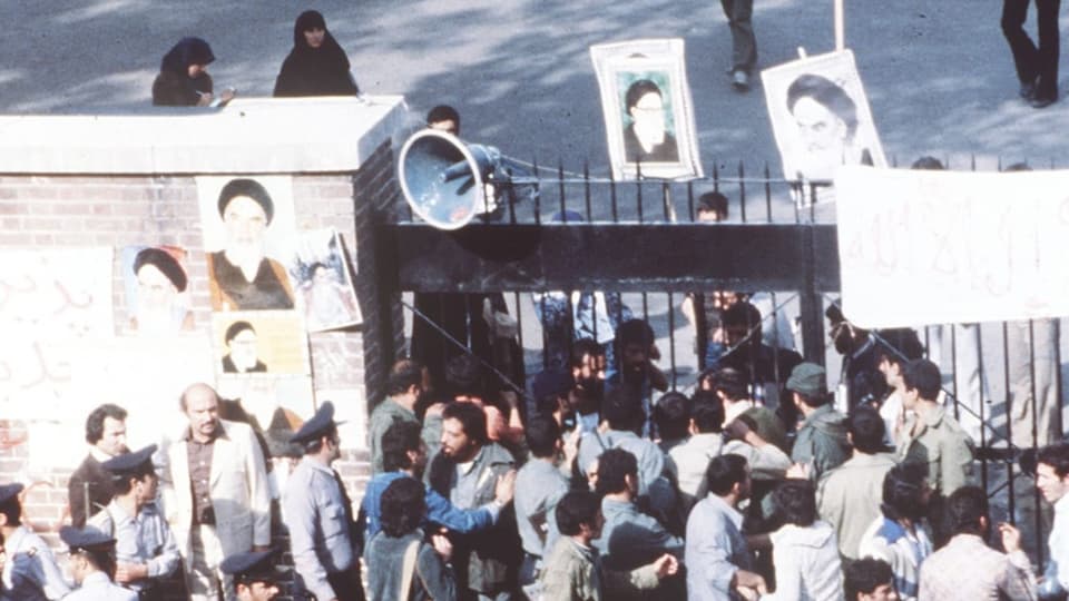 Viele Menschen stehen vor einem schwarzen hohen Tor. Dazwischen Plakate mit Schrift und Bilder eines Mannes.