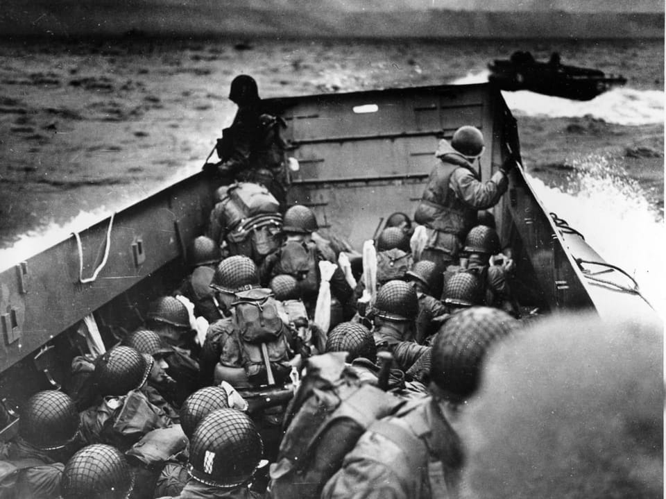 schwarzweiss-Foto Soldaten sitzen in Boot, Aufnahme leicht von oben auf ihre Helme, vor ihnen das Meer.