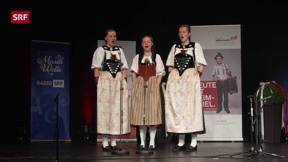 Drei Mädchen jodeln auf Bühne in Tracht.