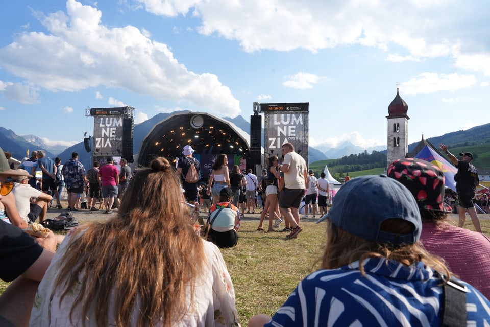 Menschenmenge bei einem Musikfestival mit Bühne und Kirchturm im Hintergrund am Open Air Lumnezia 2024.