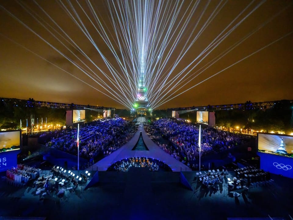 Überblick über den Veranstaltungsort Trocadero, mit dem Eiffelturm im Hintergrund und Lasern, die den Himmel erleuchten.