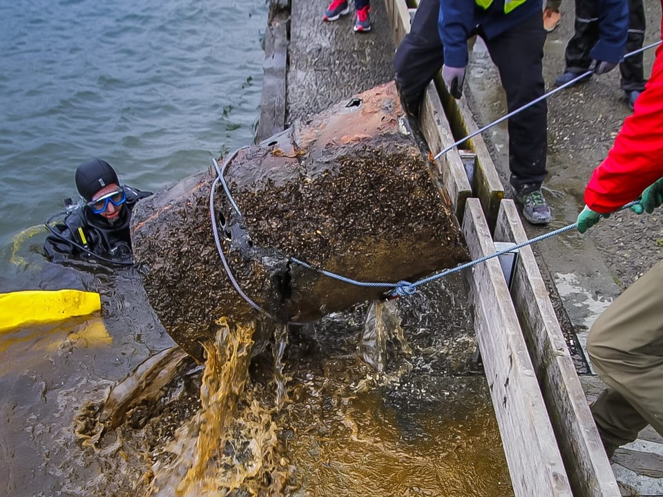 Taucher und Landarbeiter bergen ein altes Metallfass aus dem Wasser.