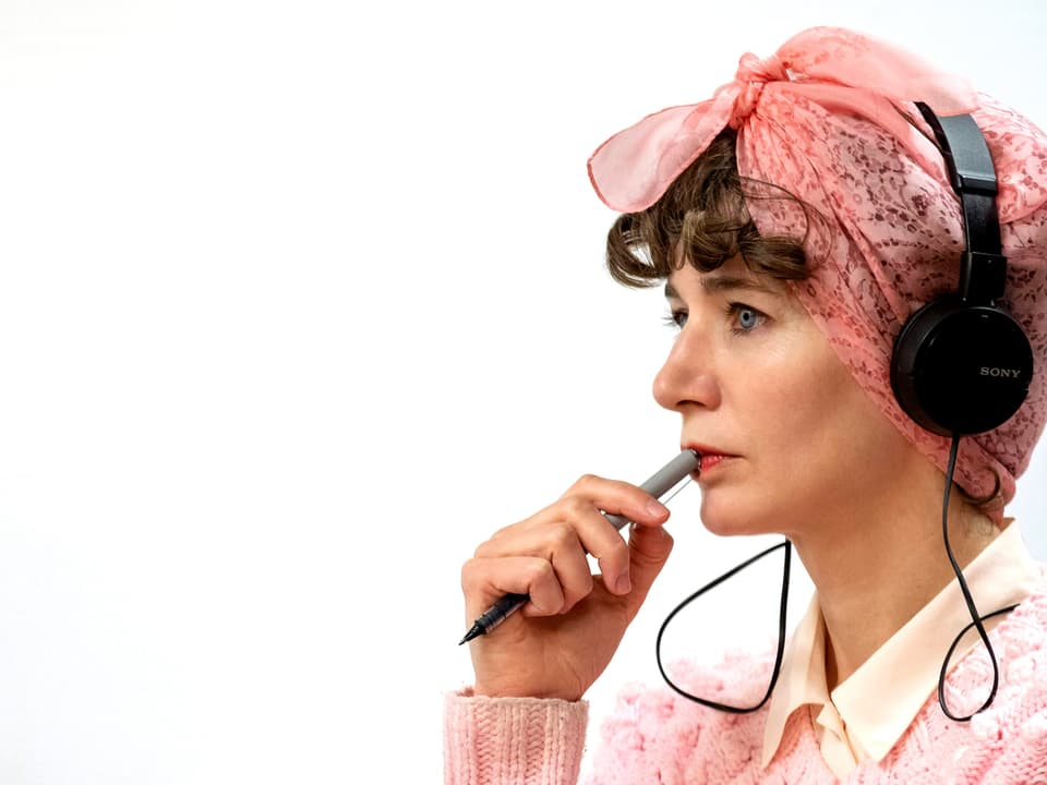 Eine Frau mit rosa Tuch um die Haare und Kopfhörern auf den Ohren in Denkerpose.