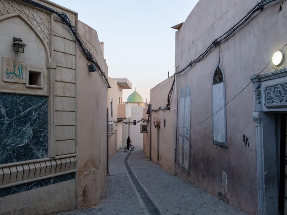Schmale Gasse zwischen hellen Gebäuden mit der Kuppel einer Moschee im Hintergrund.