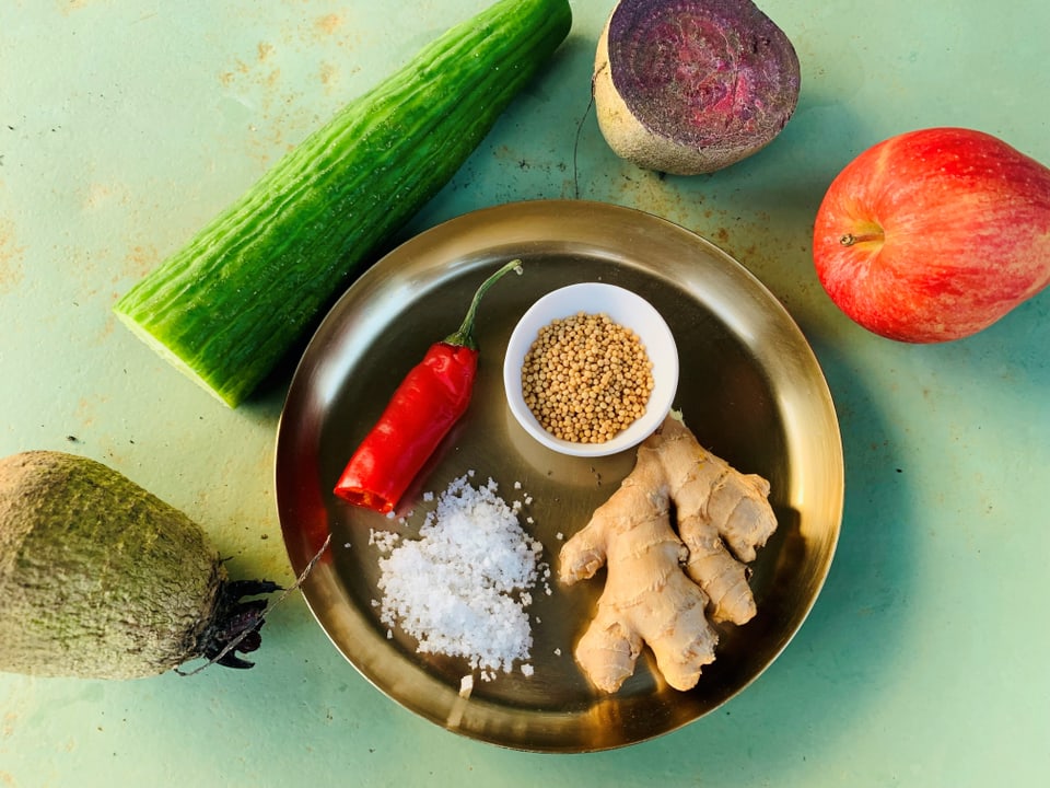 Verschiedene Zutaten auf grünem Hintergrund, darunter Gurke, Rettich, Apfel, Chili, Salz, Senfkörner, Ingwer.