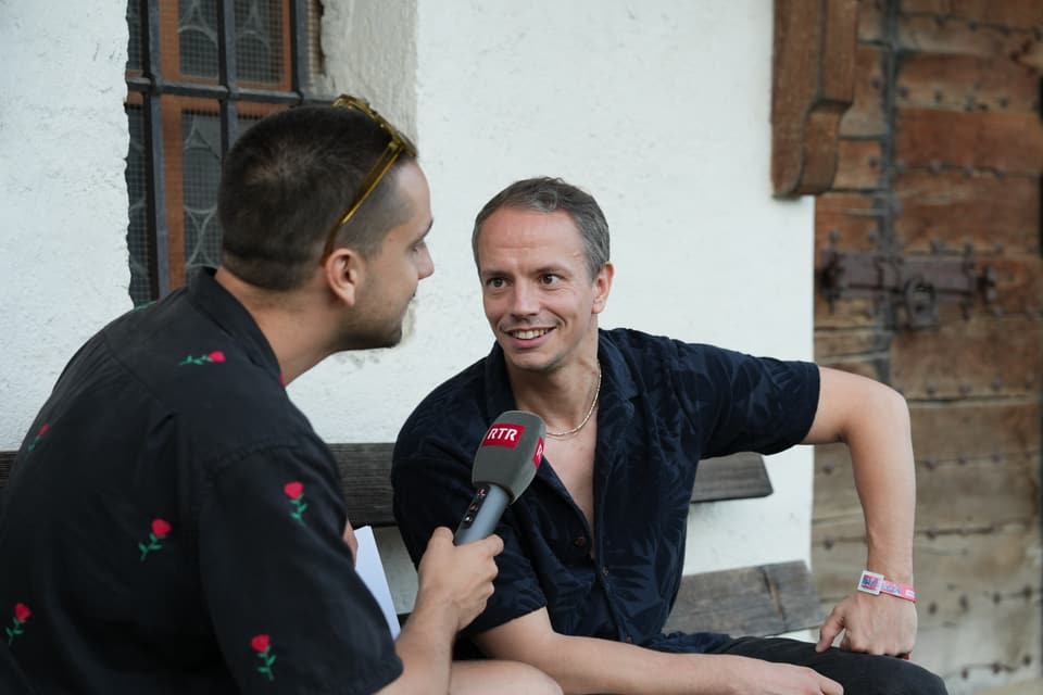 Mann interviewt anderen Mann mit Mikrofon auf einer Bank am Open Air Lumnezia 2024.