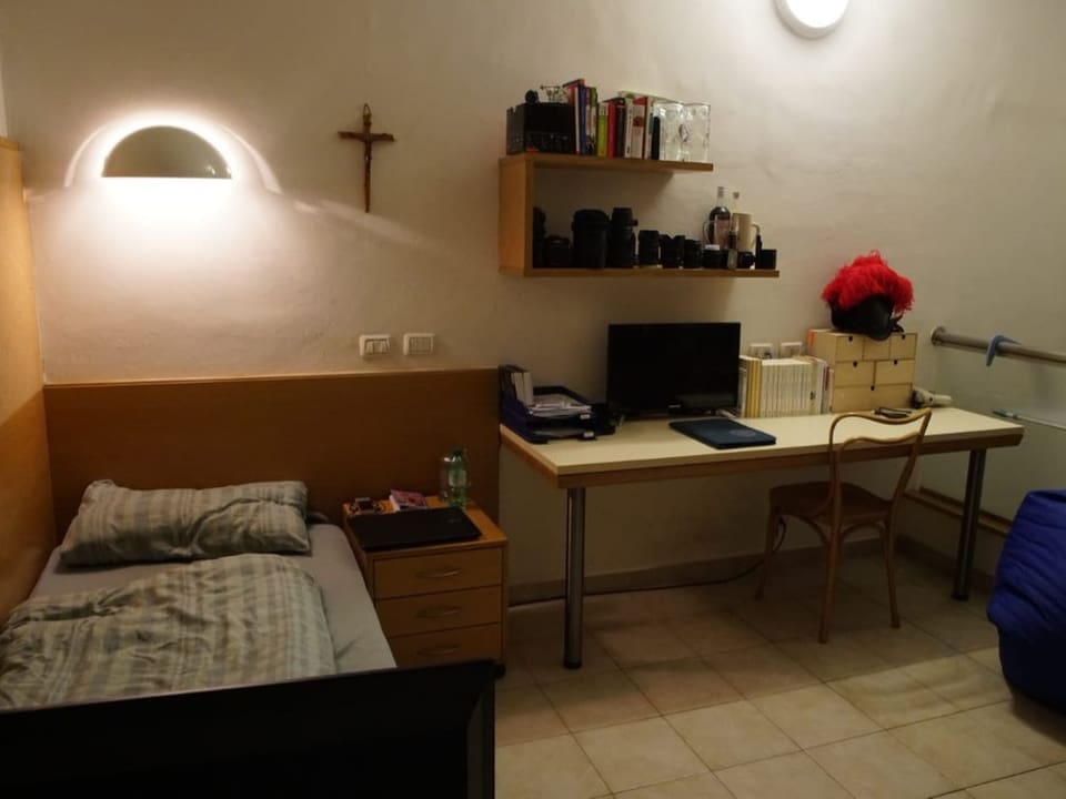 Spärlich eingerichtetes Zimmer mit Bett, Tisch, Kreuz und einigen habseligkeiten.