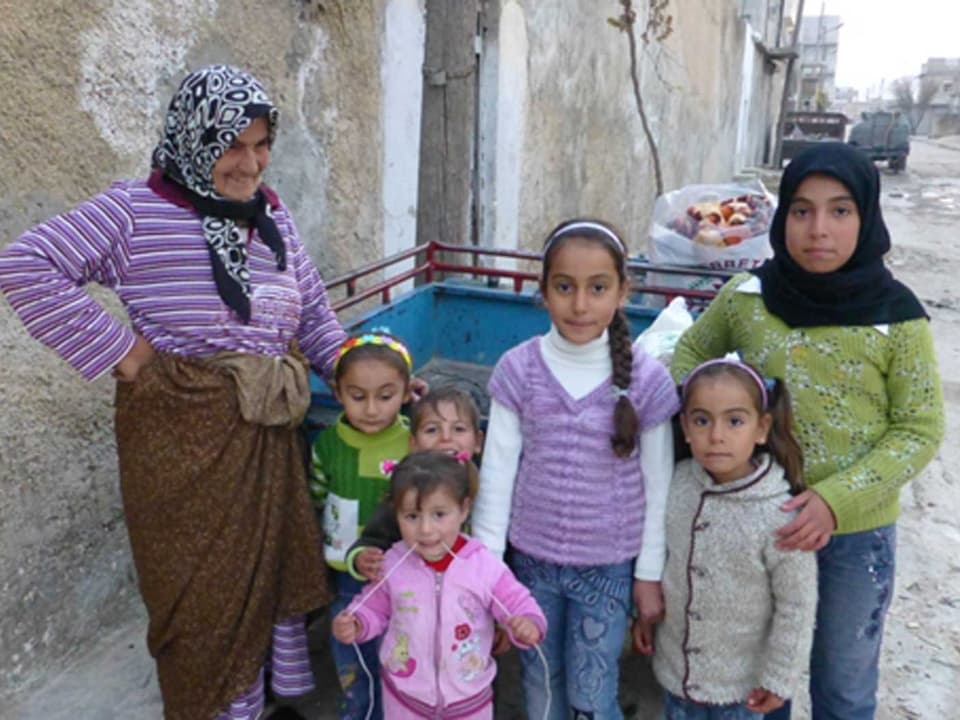 Zu sehen ist eine syrische Grossmutter mit ihren sechs Enkeln. Im Hintergrund ist eine heruntergekommene Strasse zu sehen. Ausserdem steht da ein Handkarren, in dem Lebensmittel liegen.