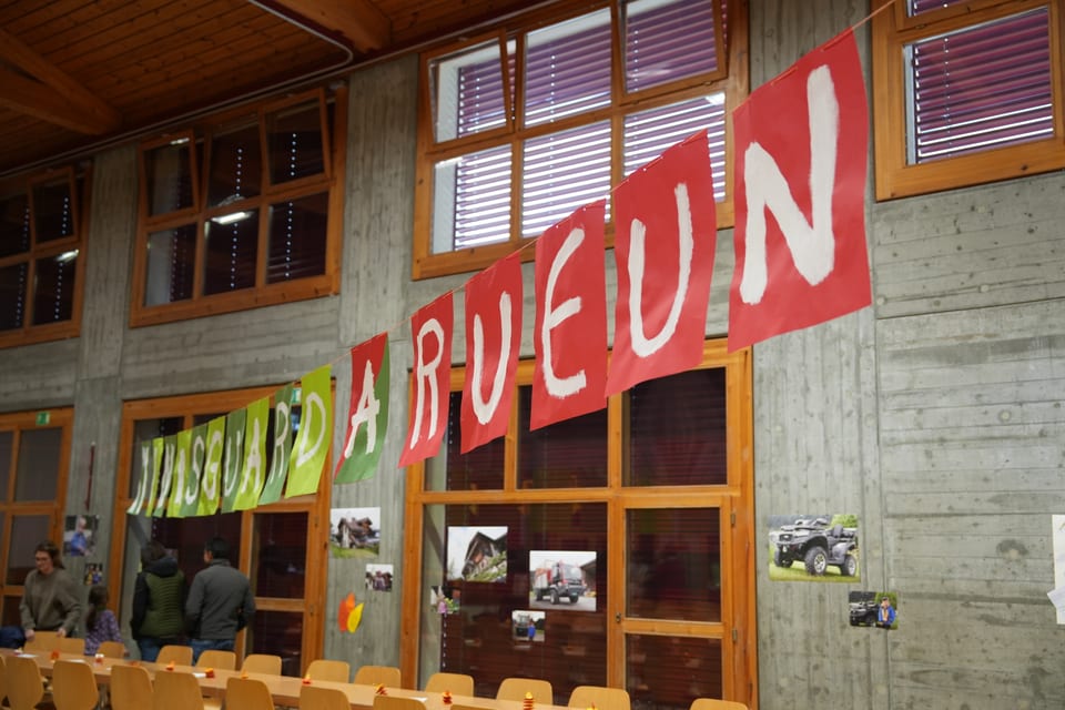 Banner mit 'Maschera a Rueun' in einem Raum mit Holzwänden.