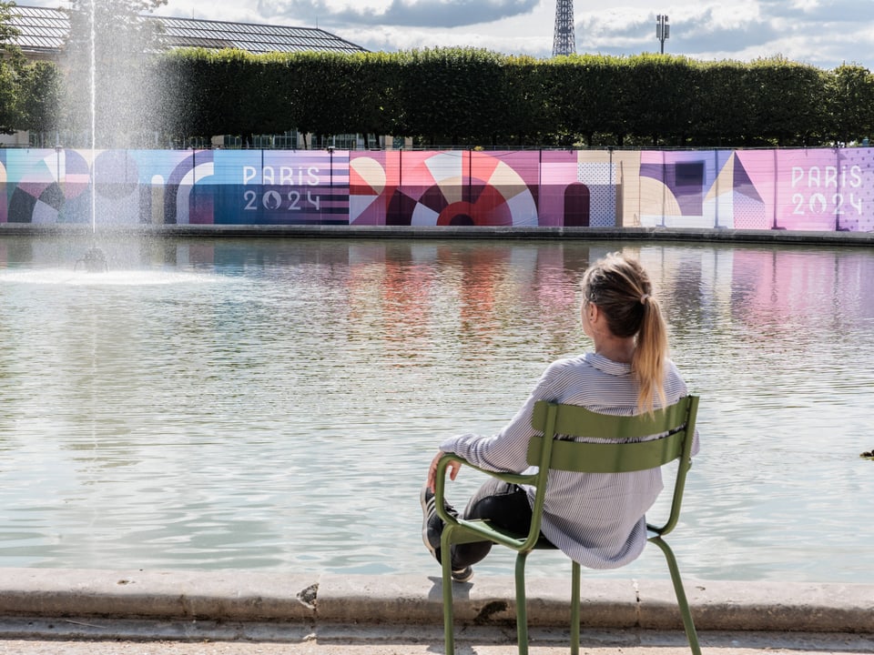 Person sitzt in einem Park vor einem Brunnen und einem Paris 2024 Plakat.